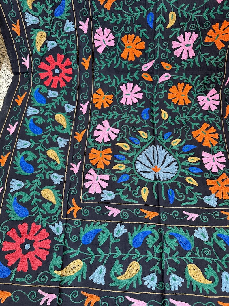 May include: Black textile with colorful floral embroidery. The design features red, orange, pink, blue, and yellow flowers and paisley shapes, with green vines and gold accents. The embroidery is detailed and covers the entire surface.
