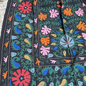May include: A dark textile with a vibrant floral embroidery design. The embroidery features red, orange, pink, blue, and yellow flowers and paisley shapes, set against a black background. Green vines and leaves complete the intricate pattern.