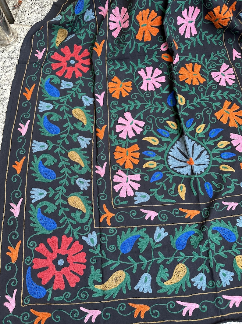 May include: A dark textile with a vibrant floral embroidery design. The embroidery features red, orange, pink, blue, and yellow flowers and paisley shapes, set against a black background. Green vines and leaves complete the intricate pattern.