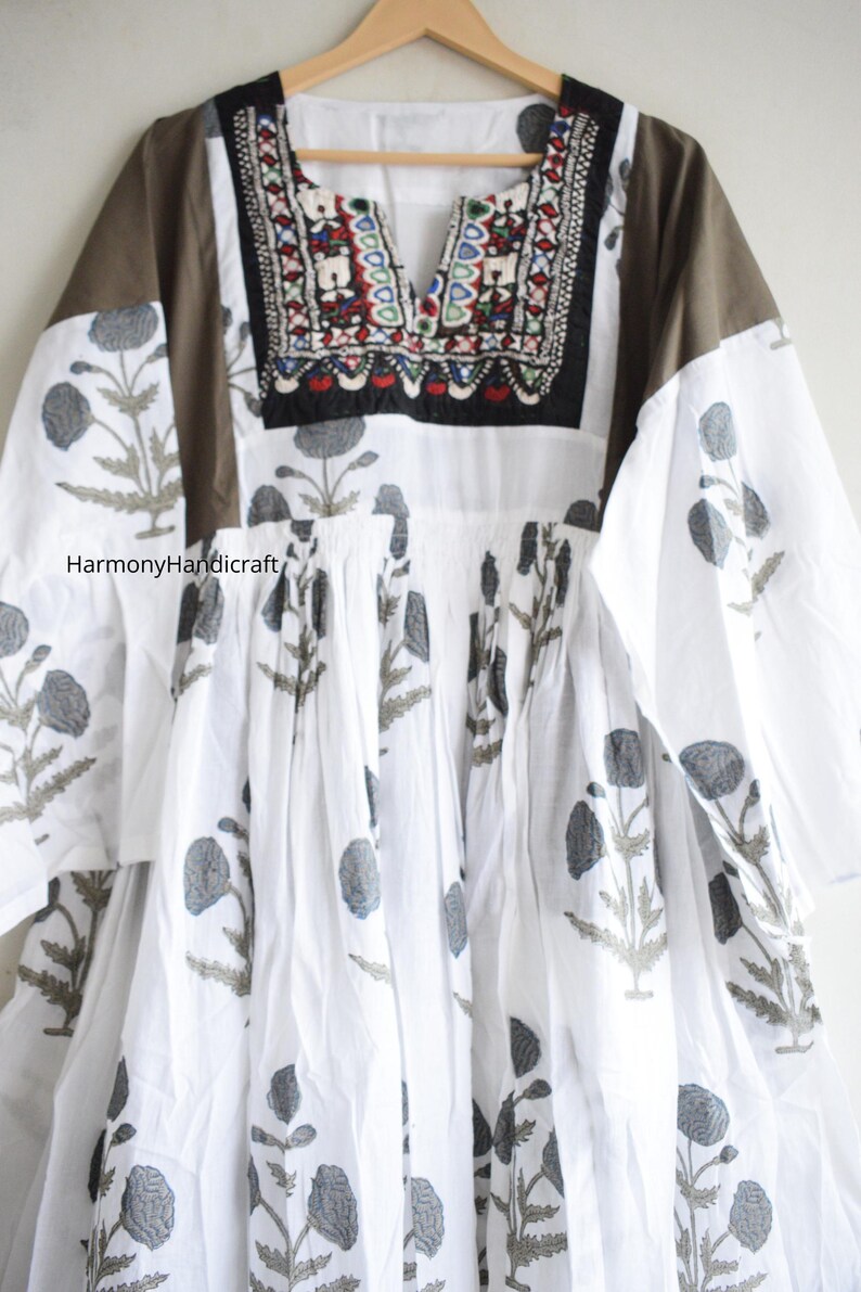Puede incluir: Vestido blanco con estampado floral gris y escote bordado. El vestido presenta mangas largas con paneles de hombros marrones y un escote en V. El texto "HarmonyHandicraft" es visible.