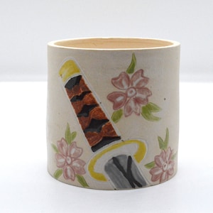 May include: A white ceramic mug with a hand-painted design of a brown and yellow katana sword with a gray blade, surrounded by pink flowers with green leaves.