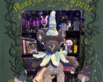 Può includere: Bambola Forest Sprite all'uncinetto fatta a mano con un design stravagante. La bambola presenta un corpo grigio scuro, un corno azzurro e bianco e un colletto verde e blu. Il testo "Fauna the Forest Sprite" è visualizzato sopra la bambola.