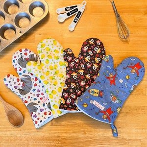 May include: Four colorful oven mitts with different patterns. One mitt has a blue background with a white star and a rocket ship. Another mitt has a black background with white skulls and flowers. A third mitt has a white background with yellow lemons and green leaves. The last mitt has a white background with red hearts and a gray seal.