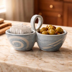 May include: A light blue ceramic serving dish with two bowls and a handle. One bowl contains packets of sugar, the other bowl is filled with green olives. The dish has a white swirl design and is sitting on a marble countertop.