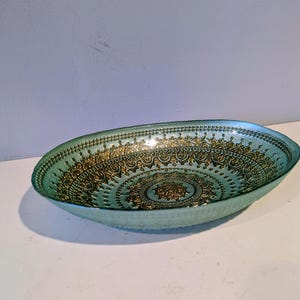 May include: Oval-shaped decorative bowl in a light teal color. The interior features an intricate gold pattern with a central medallion design. The bowl's edges are slightly raised, and the overall aesthetic is ornate.