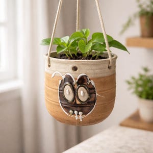 May include: A hanging ceramic planter with a whimsical face design. The planter is tan with a cream-colored rim and holds a green plant. The face is brown with white accents. The planter hangs from a natural rope.