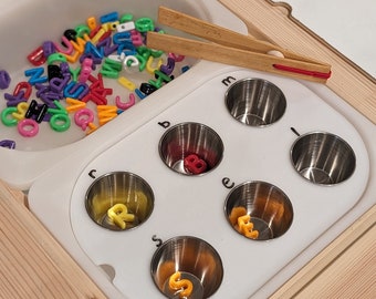 May include: A white tray with six small metal cups, each labeled with a letter of the alphabet. The cups contain colorful plastic letters. A wooden tray with a white bowl filled with colorful plastic letters is in the background. A pair of wooden tongs is on top of the tray.