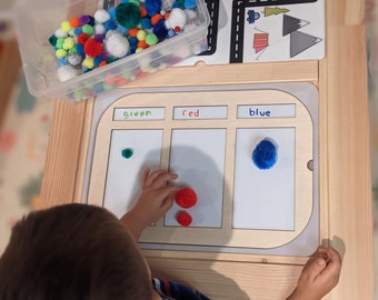 May include: A child sorts pom-poms by color. The pom-poms are in a clear container. The child is placing red pom-poms in the section labeled "red", a green pom-pom in the section labeled "green", and a blue pom-pom in the section labeled "blue".