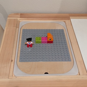 May include: A gray Lego baseplate with a small Lego figure and four Lego bricks in orange, green, pink, and red.