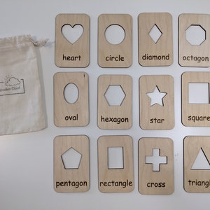 Peut inclure: Cartes de formes en bois avec des d&eacute;coupes et des &eacute;tiquettes imprim&eacute;es, comprenant c&oelig;ur, cercle, losange, etc. Un sac en toile avec le logo "The Wooden Cloud" est &agrave; gauche. Jouet &eacute;ducatif pour apprendre les formes.