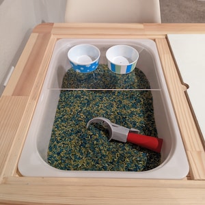 May include: A white plastic tray with a clear lid filled with green and blue rice. There are two small blue bowls and a red and gray scoop inside the tray.