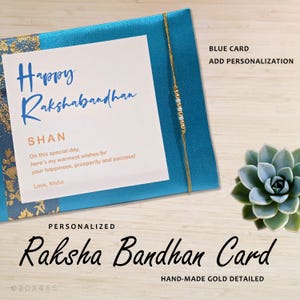 May include: A bright blue rectangular card featuring a white center panel with the text Happy Rakshabandhan SHAN and a message. A thin gold string with small white beads is wrapped around the card. Text on the card reads BLUE CARD ADD PERSONALIZATION and PERSONALIZED Raksha Bandhan Card HAND-MADE GOLD DETAILED.