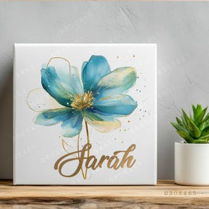 Op de afbeelding: Vierkante canvas print met een aquarel-stijl blauwe bloem met gouden accenten en de naam "Sarah" in gouden letters. De bloem heeft lichtblauwe en gouden bloemblaadjes. De achtergrond is wit.