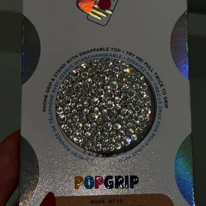 Puede incluir: Un PopSockets PopGrip con un diseño brillante similar a cristales. El embalaje incluye el logotipo de PopSockets y texto que indica que es un agarre y soporte para teléfono con una parte superior intercambiable, hecho con materiales de origen vegetal.