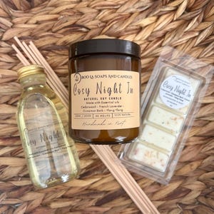 May include: A selection of aromatherapy products, including a candle, diffuser, and wax melts, presented on a woven basket. The candle jar has a brown label with the text "Cosy Night In". The diffuser bottle is clear with a gold cap.