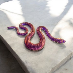 May include: A purple and copper iridescent snake sculpture with a scaly texture. The snake is curled in a figure eight shape on a grey concrete surface.
