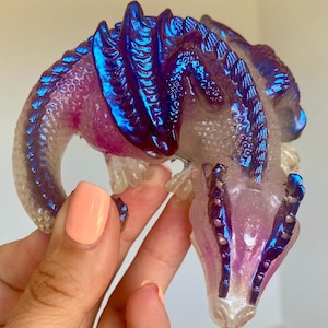May include: A small iridescent purple and blue dragon figurine with a white and purple body. The dragon has wings and a long tail.