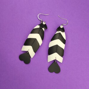 May include: A pair of black and white chevron patterned earrings with heart-shaped dangles. The earrings are made of polymer clay and have a silver hook closure.