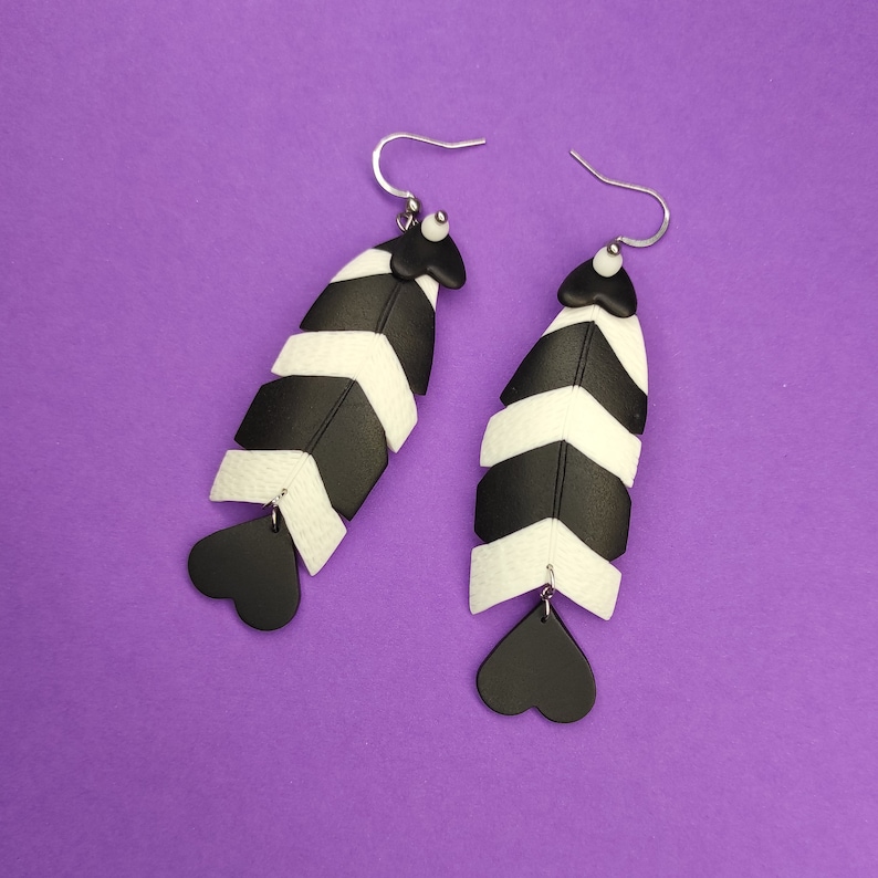 May include: A pair of black and white chevron patterned earrings with heart-shaped dangles. The earrings are made of polymer clay and have a silver hook closure.