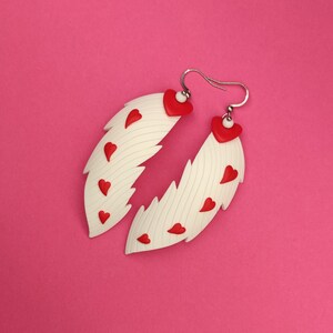 May include: White feather-shaped earrings with red heart accents. The earrings are made of polymer clay and have a textured surface.