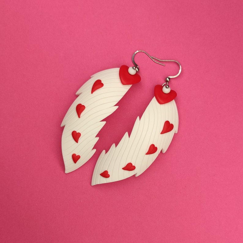 May include: White feather-shaped earrings with red heart accents. The earrings are made of polymer clay and have a textured surface.