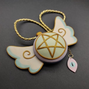 Può includere: Una collana con catena dorata con un ciondolo a forma di stella a cinque punte con contorno dorato, circondato da un cerchio verde chiaro con bordo viola. Il ciondolo è attaccato a due ali bianche con bordi dorati e un accento blu chiaro.