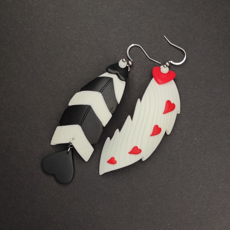 May include: A pair of earrings with a black and white chevron design and a white wing design with red hearts. The earrings are made of polymer clay and have silver hooks.