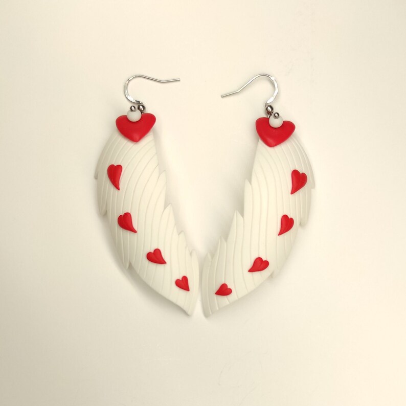 May include: A pair of white wing-shaped earrings with red heart accents. The earrings are made of polymer clay and have a silver hook.