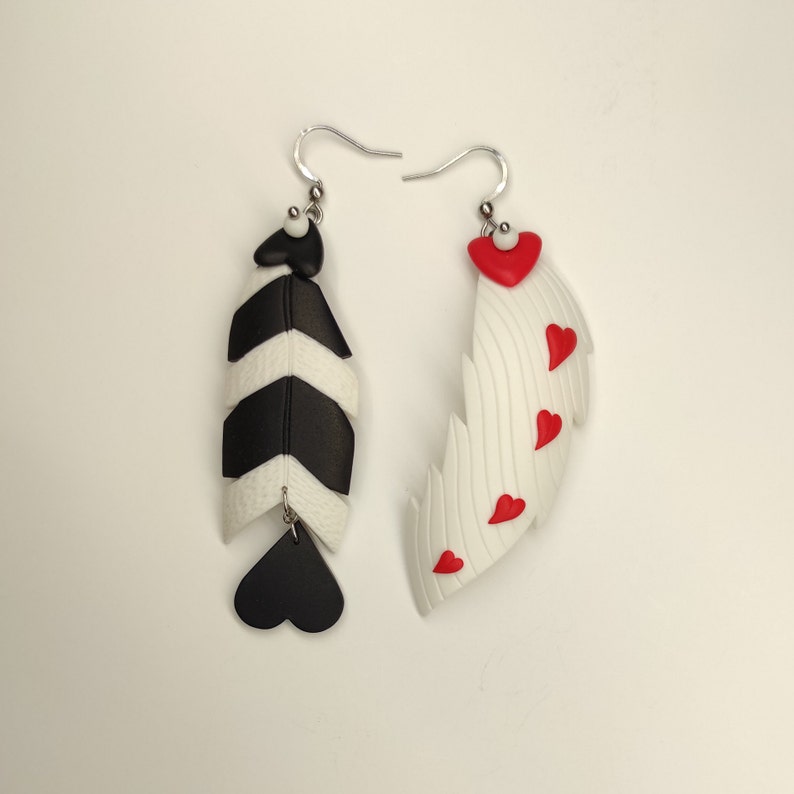 May include: A pair of dangle earrings with a black and white striped feather design on one earring and a white feather with red hearts on the other earring.