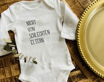 May include: A white baby onesie with long sleeves, featuring the text "NICHT VON SCHLECHTEN ELTERN" in black. The onesie is placed on a wooden surface, with a gold-colored decorative plate in the background.
