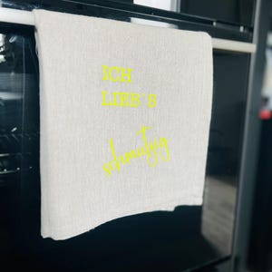 May include: A light-colored kitchen towel with the German text "ICH LIEB'S schmutzig" in yellow script. The towel is hanging on a silver bar inside a black oven. The oven's digital clock displays 08:19.