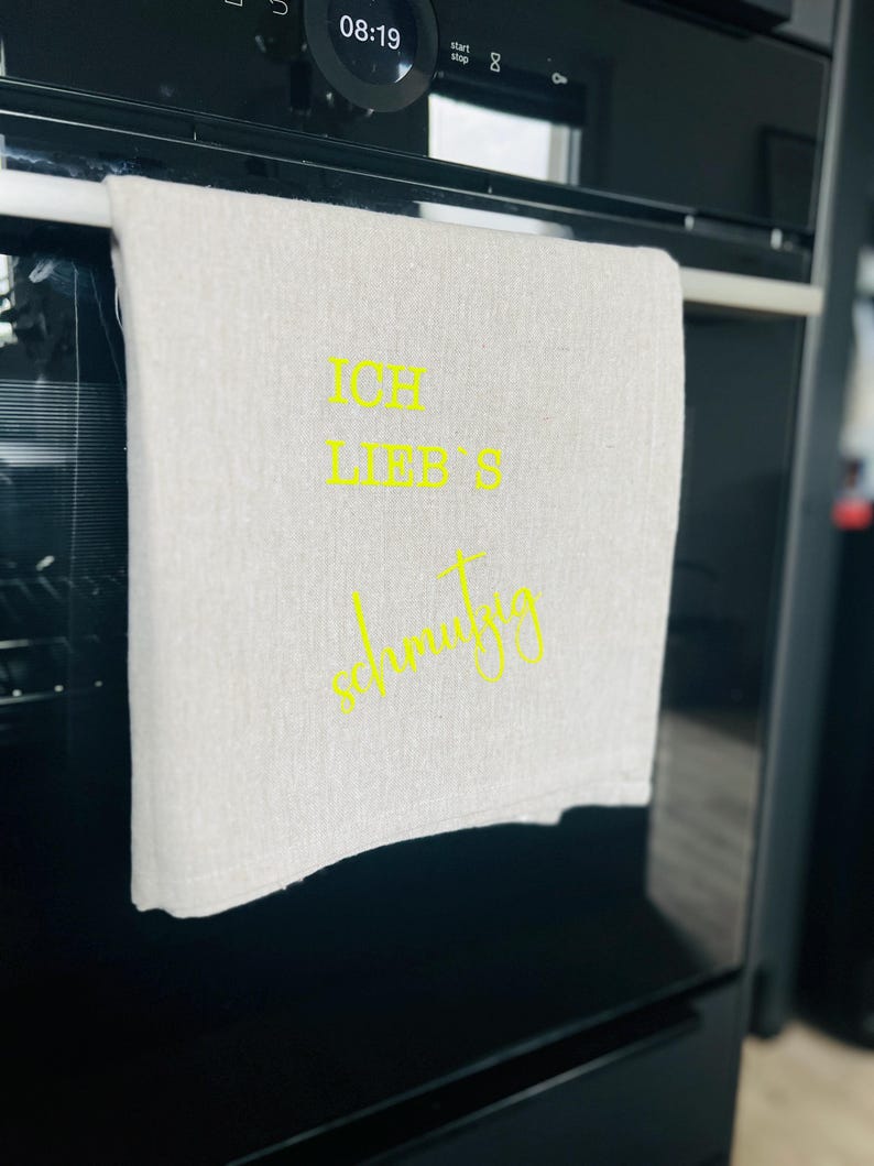 May include: A light-colored kitchen towel with the German text "ICH LIEB'S schmutzig" in yellow script. The towel is hanging on a silver bar inside a black oven. The oven's digital clock displays 08:19.