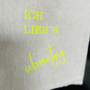 May include: A light gray linen kitchen towel with the words "ICH LIEB'S schmutzig" in bright yellow lettering. The towel is folded and hanging, with the text prominently displayed. The fabric has a textured appearance.