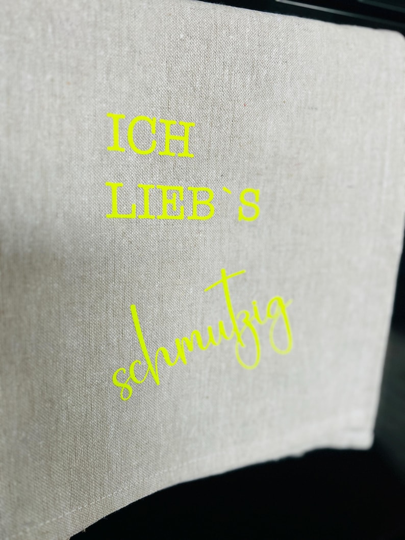 May include: A light gray linen kitchen towel with the words "ICH LIEB'S schmutzig" in bright yellow lettering. The towel is folded and hanging, with the text prominently displayed. The fabric has a textured appearance.