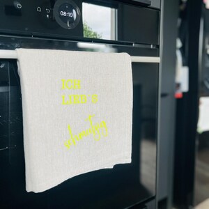 May include: A white linen kitchen towel with the text "ICH LIEB'S schmutzig" in yellow script hangs on a black oven. The oven display shows the time as 08:19. The kitchen setting has a modern aesthetic.