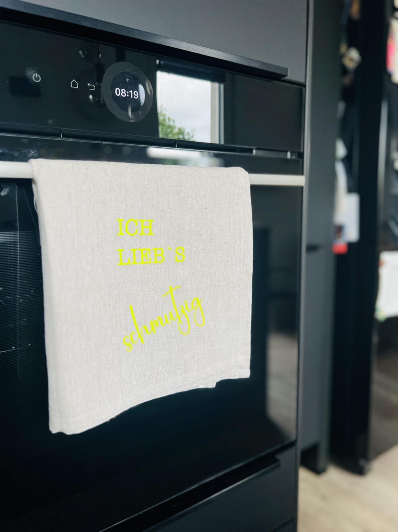 May include: A white linen kitchen towel with the text "ICH LIEB'S schmutzig" in yellow script hangs on a black oven. The oven display shows the time as 08:19. The kitchen setting has a modern aesthetic.