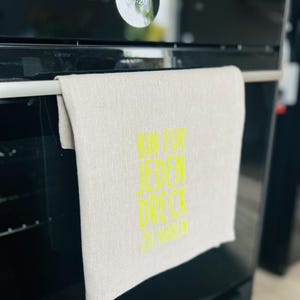 May include: A beige linen kitchen towel with the text "BIN FUR JEDEN DRECK ZU HABEN" in bright yellow. The towel hangs on a silver bar on a black oven.