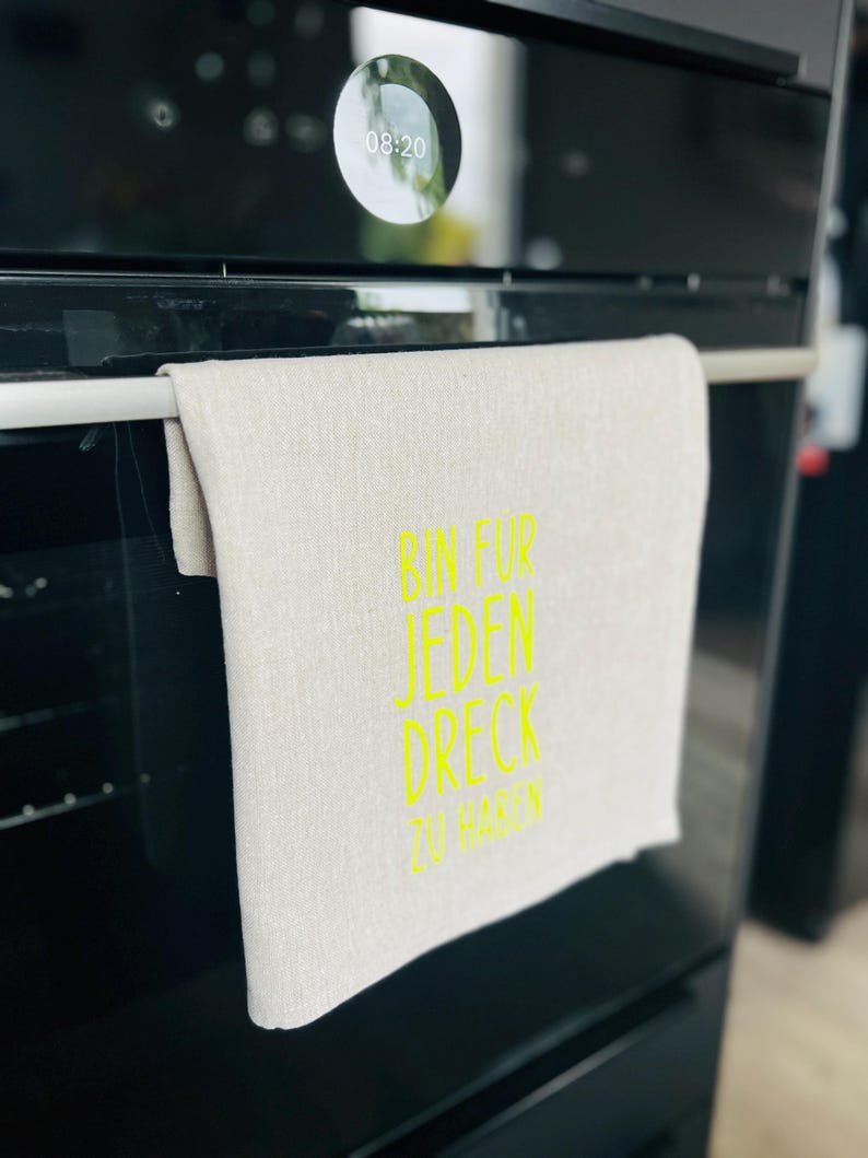 May include: A beige linen kitchen towel with the text "BIN FUR JEDEN DRECK ZU HABEN" in bright yellow. The towel hangs on a silver bar on a black oven.
