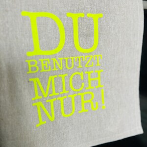 May include: A light gray linen kitchen towel with the words "DU BENUTZT MICH NUR!" in bright yellow. The towel is folded and hanging, with the text prominently displayed.