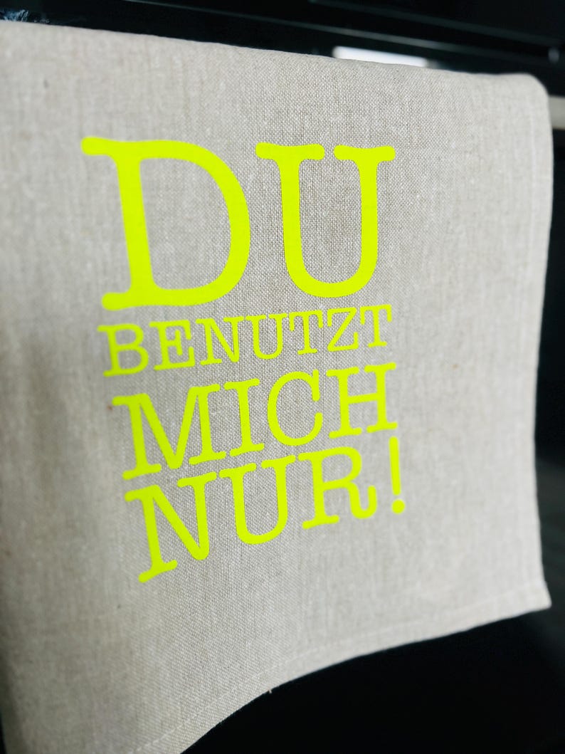 May include: A light gray linen kitchen towel with the words "DU BENUTZT MICH NUR!" in bright yellow. The towel is folded and hanging, with the text prominently displayed.