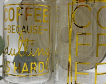 May include: Two clear glass mugs with gold lettering. The mug on the left says "Coffee -Because- Adulting is Hard". The mug on the right says "Coffee". Both mugs are sitting on a wooden surface with coffee beans scattered around them.