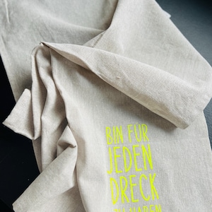 May include: A beige linen kitchen towel with the text "BIN FUR JEDEN DRECK ZU HABEN" in bright yellow. The towel is folded and draped, showcasing its texture and design. The text is centered and easy to read.