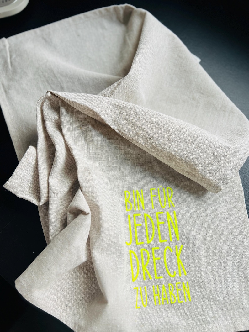 May include: A beige linen kitchen towel with the text "BIN FUR JEDEN DRECK ZU HABEN" in bright yellow. The towel is folded and draped, showcasing its texture and design. The text is centered and easy to read.