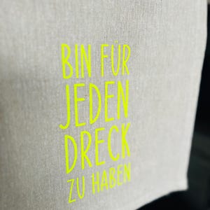 May include: A light gray linen towel with the neon green text "BIN FUR JEDEN DRECK ZU HABEN". The towel is folded over, with the text visible on the front. The background is dark.