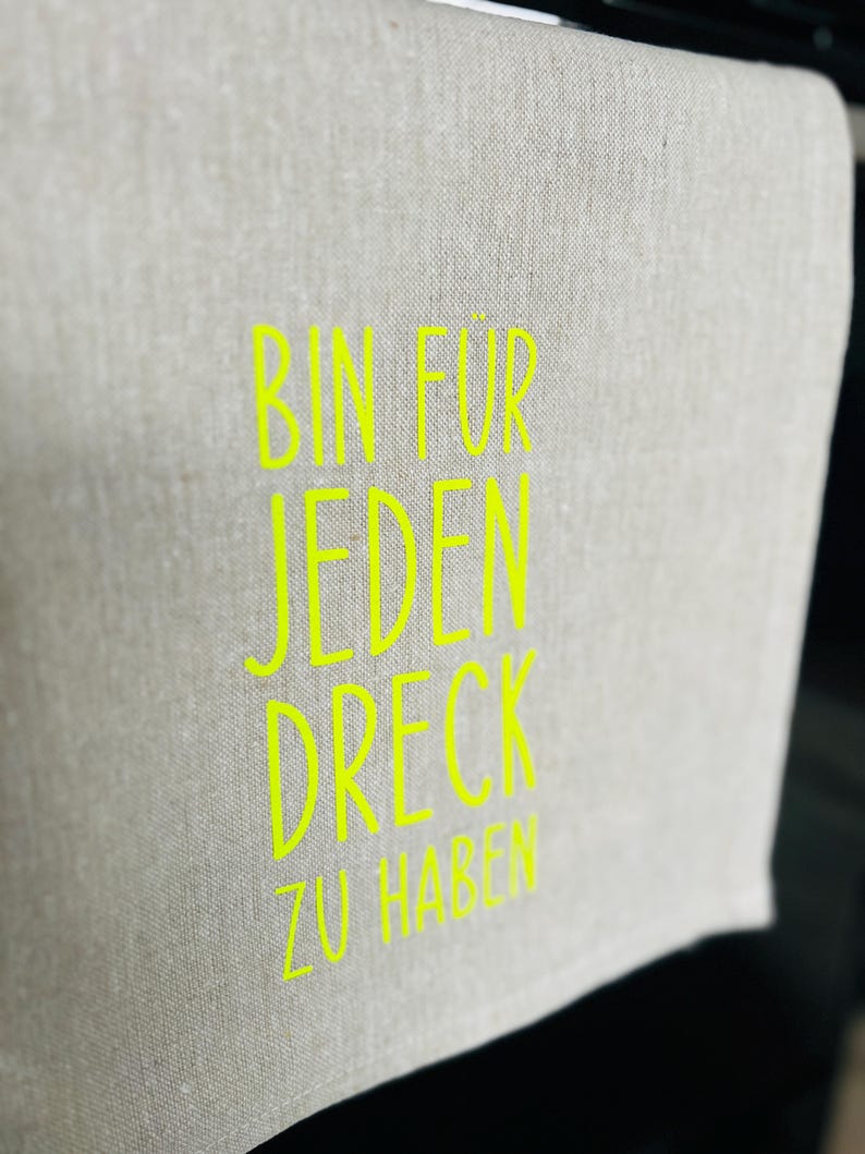 May include: A light gray linen towel with the neon green text "BIN FUR JEDEN DRECK ZU HABEN". The towel is folded over, with the text visible on the front. The background is dark.