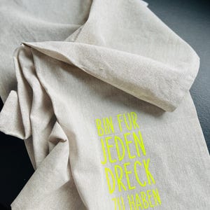 May include: A folded, beige linen towel with the neon green text "BIN FUR JEDEN DRECK ZU HABEN". The towel is made of a soft, textured fabric and is folded over itself.