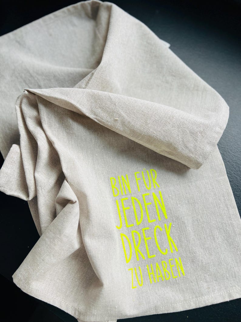 May include: A folded, beige linen towel with the neon green text "BIN FUR JEDEN DRECK ZU HABEN". The towel is made of a soft, textured fabric and is folded over itself.