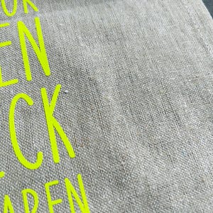 May include: Close-up of a beige linen fabric with neon yellow text. The fabric has a textured appearance. The text is partially visible, with the letters "UR", "EN", "CK", and "ABEN" in a bold, sans-serif font.