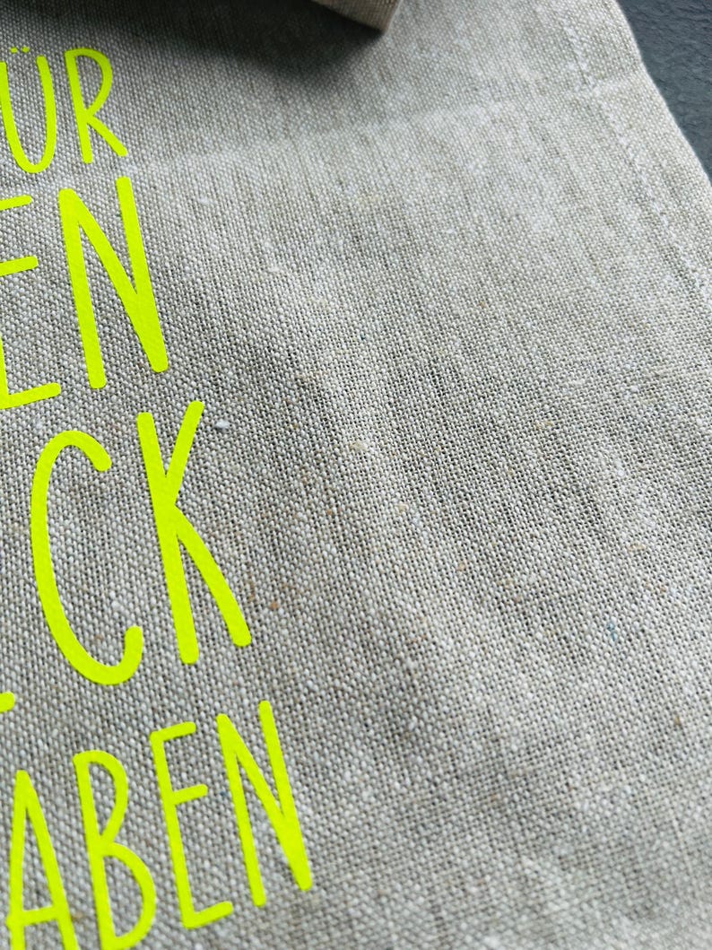 May include: Close-up of a beige linen fabric with neon yellow text. The fabric has a textured appearance. The text is partially visible, with the letters "UR", "EN", "CK", and "ABEN" in a bold, sans-serif font.