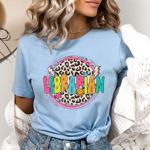 May include: A light blue t-shirt with a colorful graphic of the word "LIBRARIAN" in a circle with a leopard print background. The graphic also includes lightning bolts and stars.